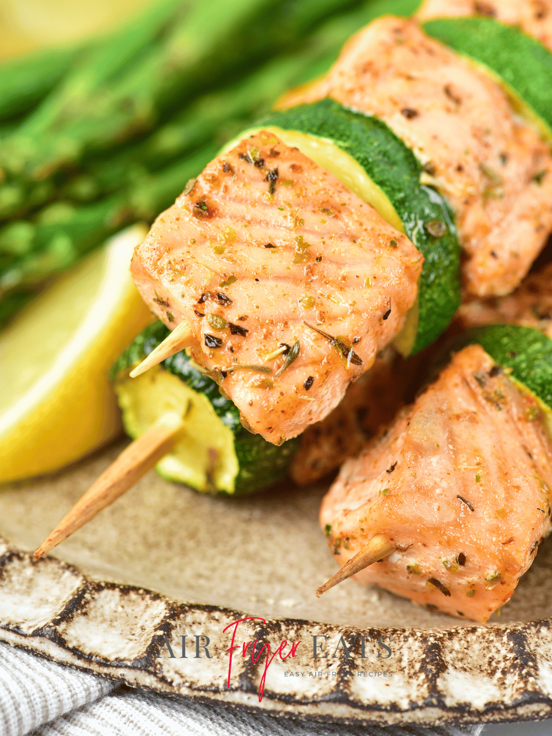 A close-up shot of air fryer salmon skewers. The salmon bites are crispy and pink, with flakes of seasonings on them. There are asparagus and lemon wedges in the background.