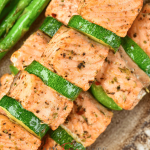 A close-up shot of a salmon skewer. The salmon is arranged between slices of zucchini and green bell peppers. They are plated alongside asparagus, a great side for seafood.