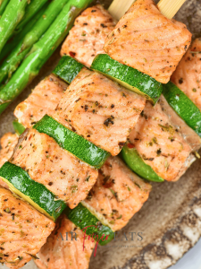 A close-up shot of a salmon skewer. The salmon is arranged between slices of zucchini and green bell peppers. They are plated alongside asparagus, a great side for seafood.