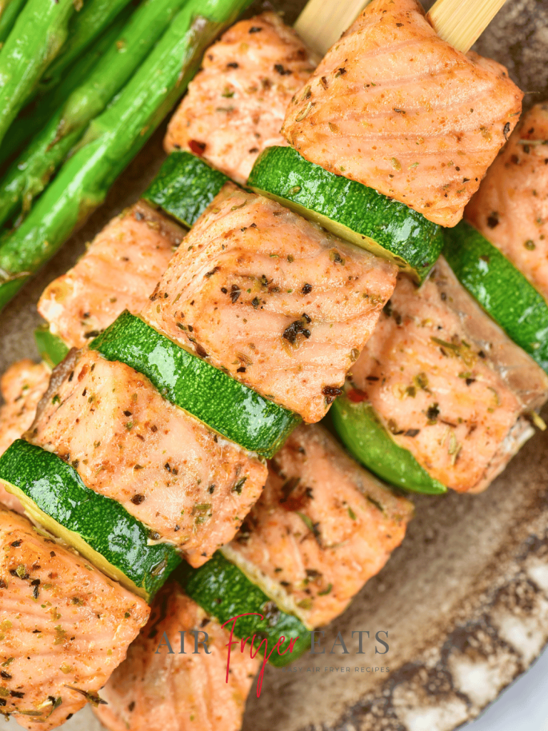 A close-up shot of a salmon skewer. The salmon is arranged between slices of zucchini and green bell peppers. They are plated alongside asparagus, a great side for seafood.