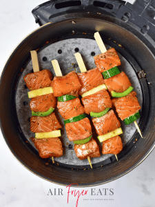 Salmon pieces and veggie pieces are arranged on wooden skewers and placed in an air fryer basket, ready to be cooked.