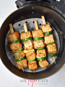 Four cooked air fryer salmon skewers are arranged neatly in a round, black air fryer basket on top of an air fryer liner. The salmon skewers are orange and golden and covered with green seasoning flakes.