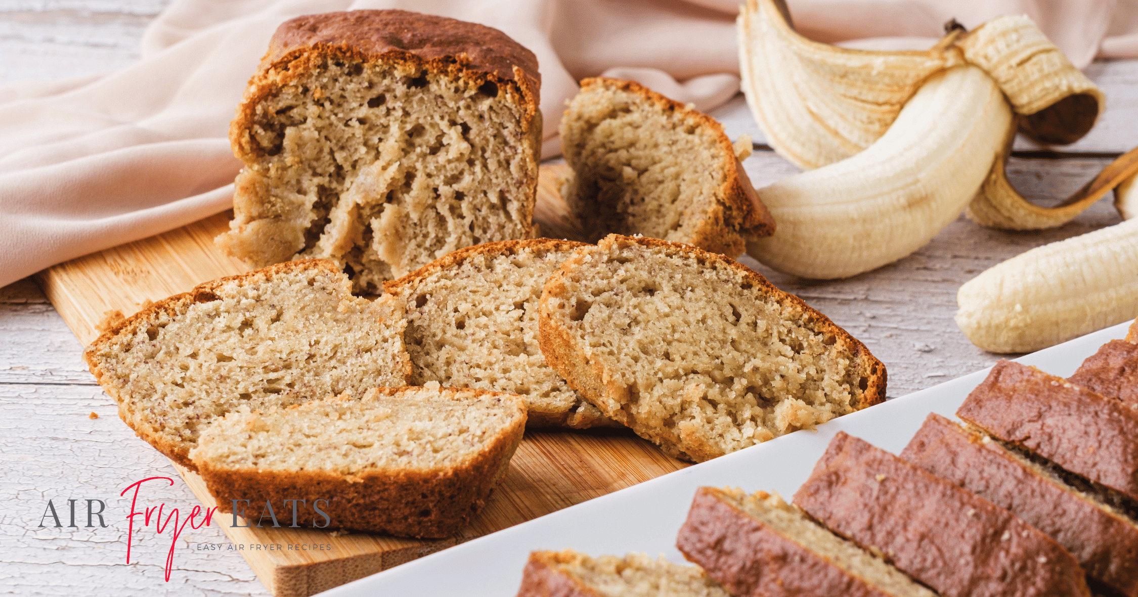 A close-up shot of a loaf of banana bread made in the air fryer. There are freshly cut slices of bread and a few unpeeled bananas in the background. The bread is fluffy, moist, and sweet.