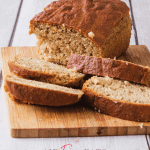A loaf of air fryer banana bread sits on a wooden cutting board, there are a few slices of banana bread laying on the cutting board as well. The top is golden brown and crispy and the inside is moist and fluffy.