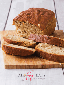 A loaf of air fryer banana bread sits on a wooden cutting board, there are a few slices of banana bread laying on the cutting board as well. The top is golden brown and crispy and the inside is moist and fluffy.
