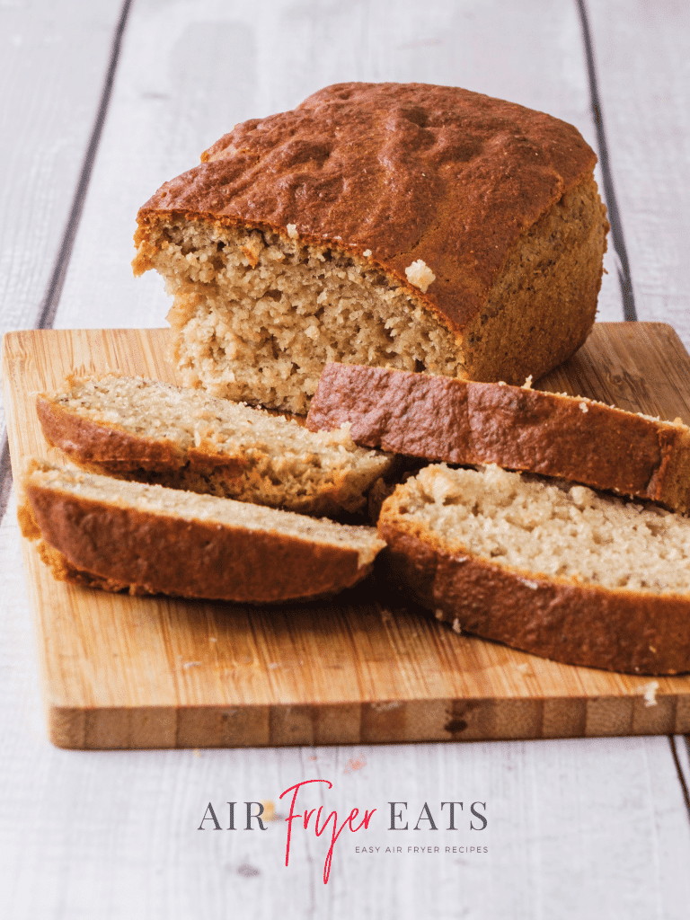 A loaf of air fryer banana bread sits on a wooden cutting board, there are a few slices of banana bread laying on the cutting board as well. The top is golden brown and crispy and the inside is moist and fluffy.