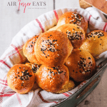 A tin can with a red and white kitchen towel holds several small air fryer bagel bites. They are golden brown and covered with white and black sesame seeds. The tops of the bagel bites are shiny and soft.