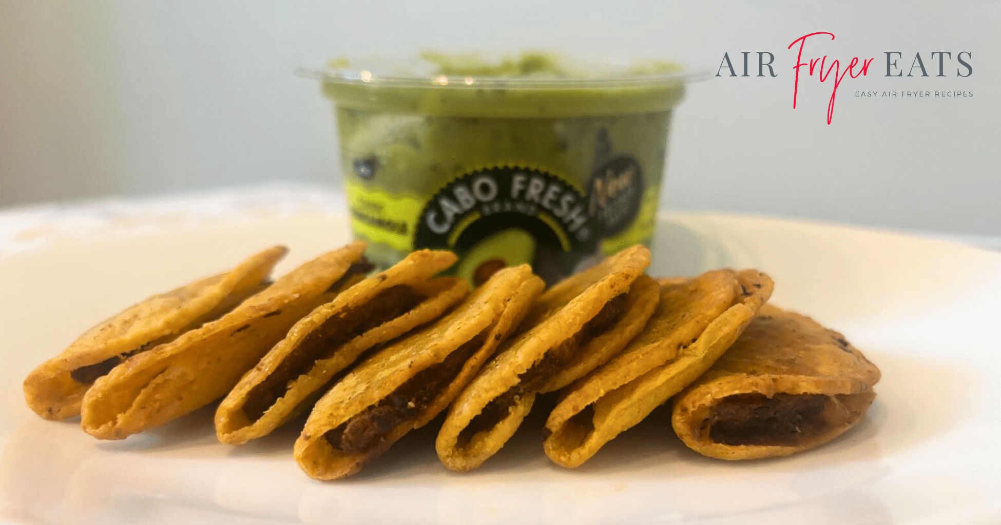 A close-up shot of seven jose ole mini tacos made in the air fryer on a plate. In the background there is a container of guacamole for dipping the mini tacos in.
