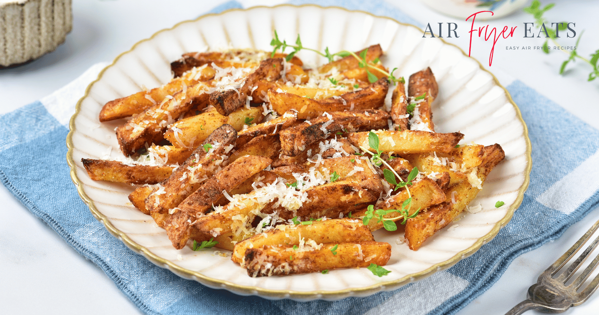 A white plate with gold edges is filled with crispy golden brown garlic parmesan fries made in the air fryer. The plate is set atop a blue and white kitchen towel and there is a silver fork nearby.