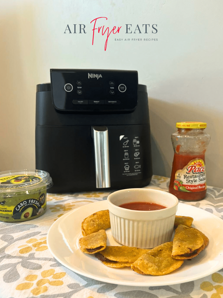 A plate of cooked jose ole mini tacos air fryer are arranged on a kitchen table next to guacamole, Pace salsa, and a large, black Ninja air fryer. The mini tacos are crispy and bite-sized.