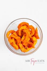 A glass bowl is full of bright orange sliced pumpkin pieces covered with red seasonings.