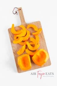 A small pumpkin is peeled and sliced in half on a wooden cutting board. There are two larger pumpkin pieces and many small slices of pumpkin that will be used to make air fryer pumpkin fries.