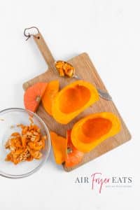 A small orange pumpkin is sliced in half and the seeds are removed and placed in a glass bowl alongside the cutting board. The top and bottom of the pumpkin has been cut off and lies alongside the pumpkin pieces.