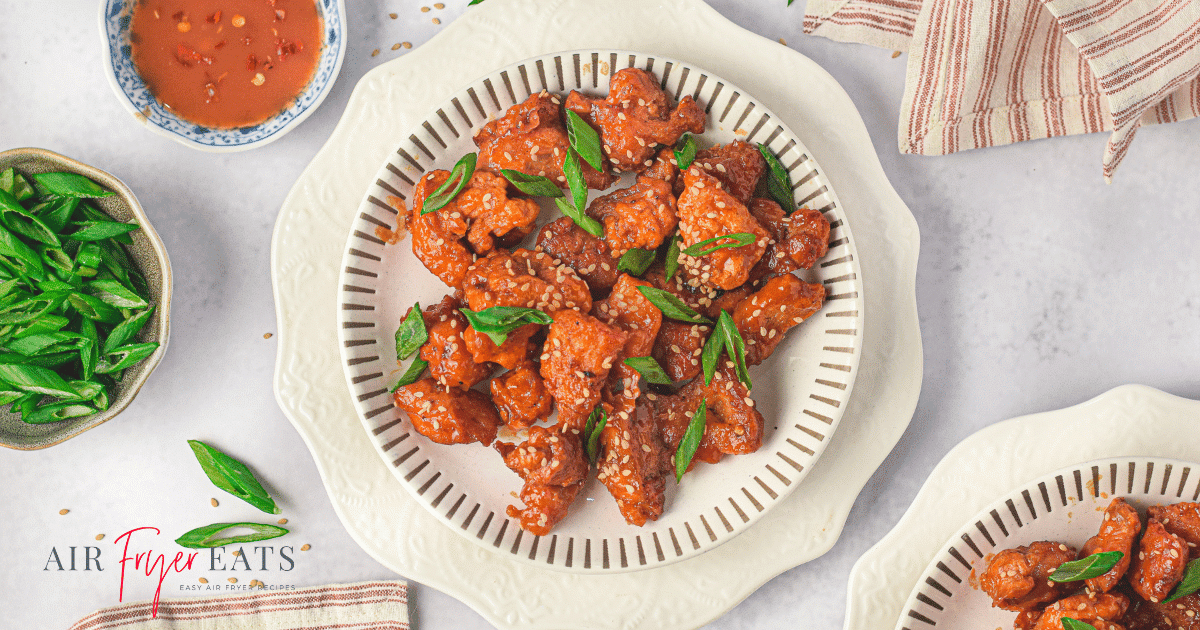 Air fryer Asian chicken is plated on a white plate. The chicken pieces are a bright orange red color and are covered in green onion slices and sesame seeds. There is a kitchen towel nearby and a bowl of spicy red sauce.