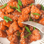 A close-up shot of a plate of air fryer Asian chicken. A fork is piercing the middle piece of breaded chicken and all the chicken pieces are garnished with green onions and sesame seeds.