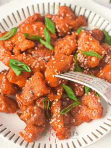 A close-up shot of a plate of air fryer Asian chicken. A fork is piercing the middle piece of breaded chicken and all the chicken pieces are garnished with green onions and sesame seeds.