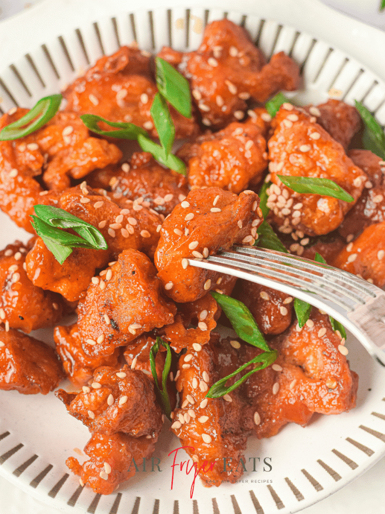 A close-up shot of a plate of air fryer Asian chicken. A fork is piercing the middle piece of breaded chicken and all the chicken pieces are garnished with green onions and sesame seeds.