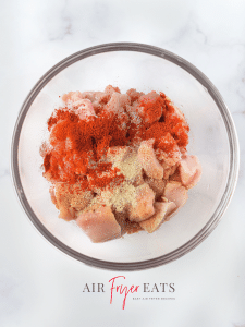 A glass bowl is full of cubed raw chicken breasts that are covered in red, brown, and white seasonings.