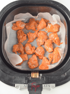 About a dozen seasoned Asian chicken pieces are arranged neatly on a parchment paper inside an air fryer basket. They are covered in a red, wet batter.