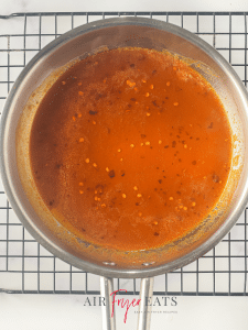 A metal saucepan is full of a orange sauce with red chili flakes inside. The saucepan is placed on a wire rack on a kitchen counter.