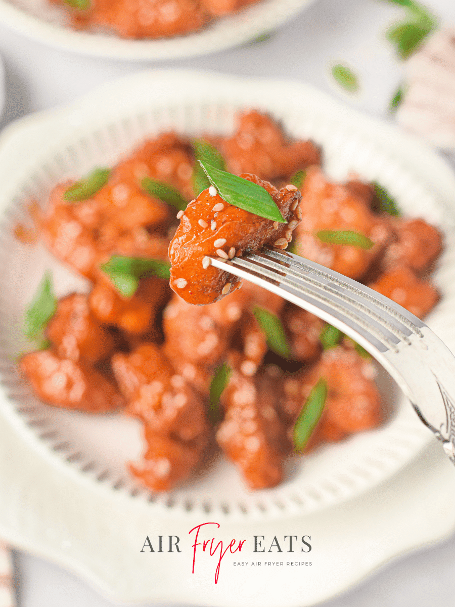 A silver fork holds a piece of air fryer Asian chicken. The chicken bite is crispy and coated in a sticky red sauce. The chicken pieces in the background are plated and topped with sesame seeds and green onion slices.