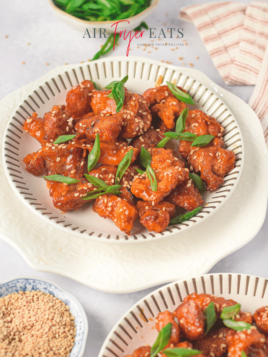 A plate of air fryer sticky chicken. The chicken pieces are garnished with green onion and sesame seeds. There is a kitchen towel in the background. Another bowl of air fryer sticky chicken is in the foreground, along with a small bowl of sesame seeds.