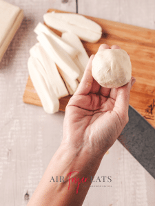 A home chef rolls a mozzarella dough into a neat ball. In the background, there are string cheese sticks and a wooden cutting board.