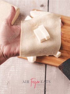 A home chef places two small pieces of mozzarella cheese in the center of a Pillsbury crescent roll dough square.