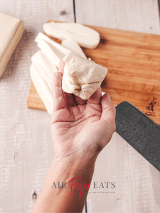 A home chef folds crescent roll dough over pieces of mozzarella string cheese to form a dough ball. In the background, there are string cheese sticks and a cutting board.