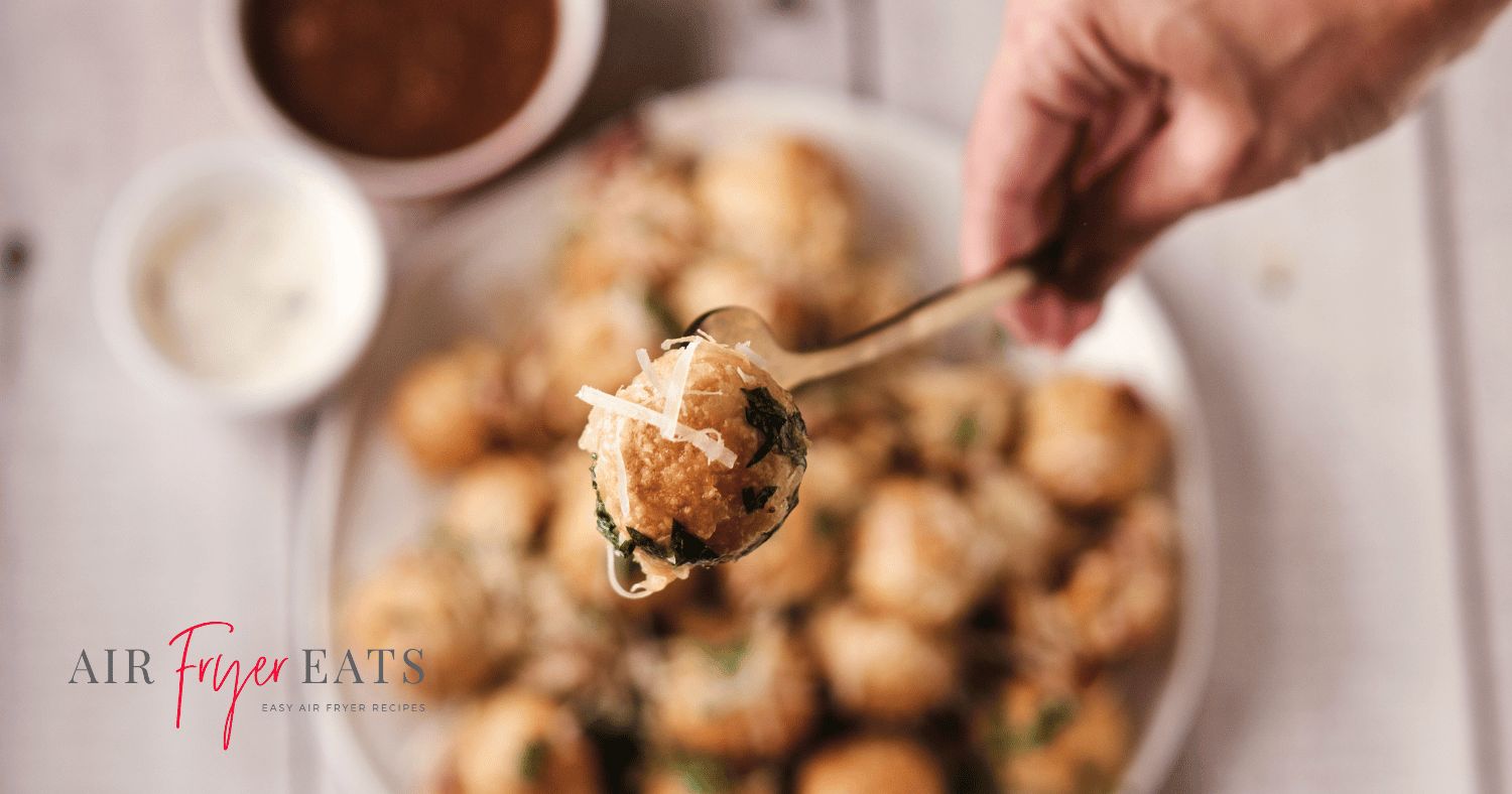 A close-up shot of a single air fryer mozzarella bite covered with parmesan cheese and parsley. In the background, there is a plate full of mozzarella bites and two bowls with dipping sauces.