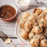 A large white plate is full of crispy air fryer mozzarella bites. They are covered in freshly-grated parmesan cheese and parsley. Nearby, there are marinara and ranch dipping sauces.