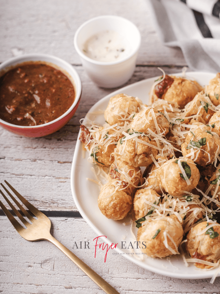 A large white plate is full of crispy air fryer mozzarella bites. They are covered in freshly-grated parmesan cheese and parsley. Nearby, there are marinara and ranch dipping sauces.