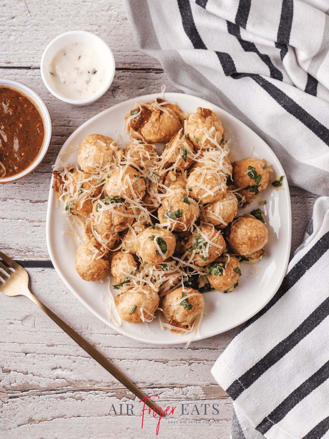 A white plate is full of air fryer mozzarella bites covered in fresh parsley and parmesan cheese. Nearby, there is a black and white striped kitchen towel, a gold fork, and two small bowls filled with marinara and ranch dipping sauces.