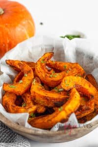 A close-up shot of crunchy air fryer pumpkin fries. They are dark orange and slightly browned on the edges. The fries lie in a brown bowl and are covered in freshly chopped parsley. There is a large orange pumpkin in the background.