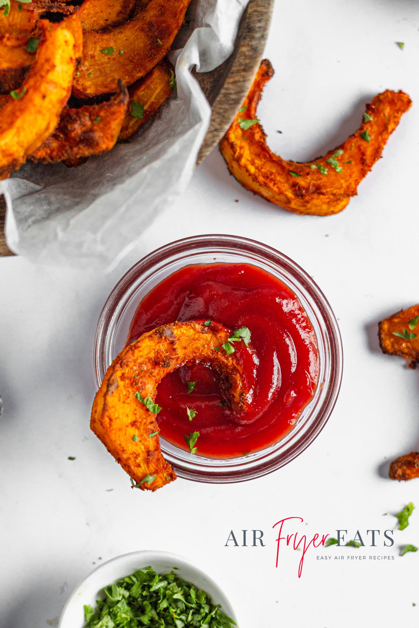 An air fryer pumpkin fry is dipped in a small glass bowl of ketchup. Nearby, a bowl of air fryer pumpkin fries, and on the white kitchen counter there are a few pumpkin fries with parsley.